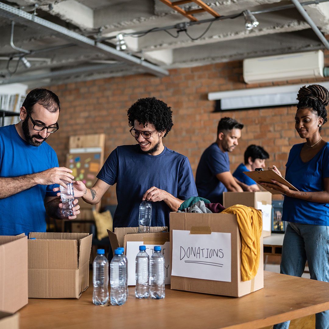 Volunteers arranging donations in a community charity donation center | UW Comal and Guadalupe Counties Group of volunteers in blue shirts packing donated items into boxes labeled DONATIONS in a brick-walled room.