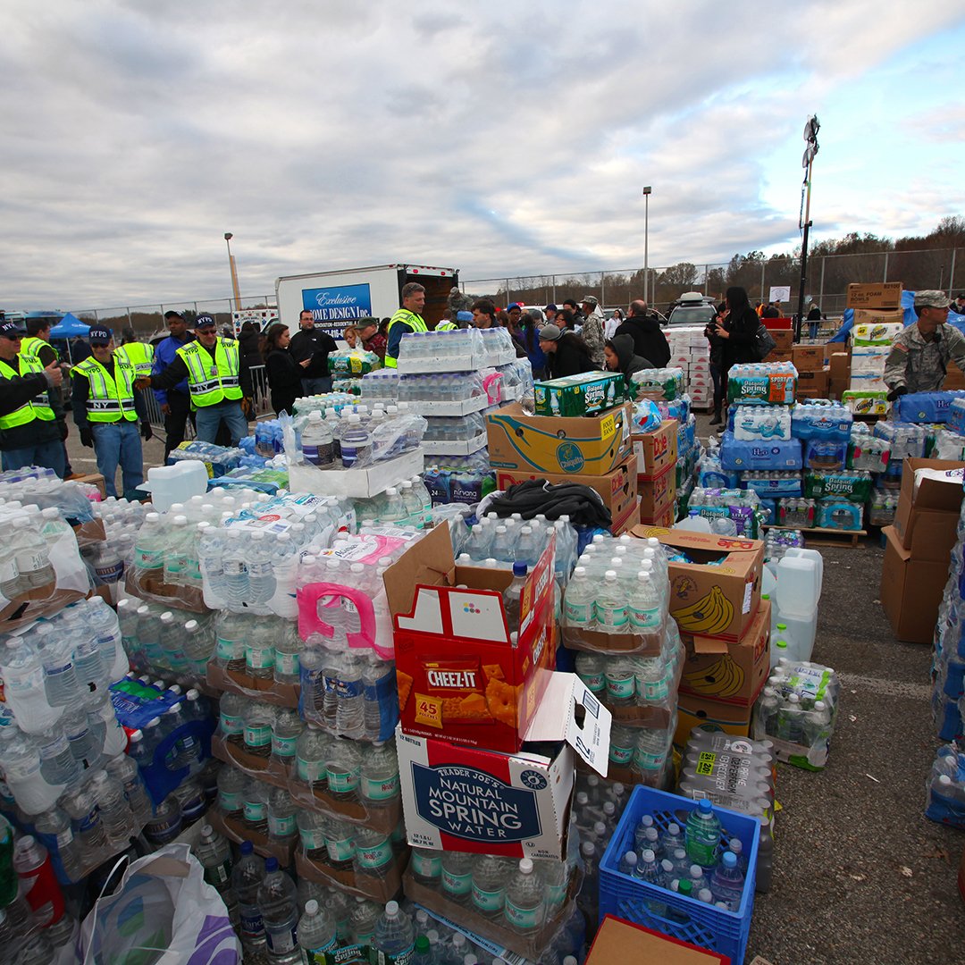 | UW Comal and Guadalupe Counties Large outdoor distribution scene with volunteers in neon safety vests organizing bottled water and boxes of supplies in a parking lot.