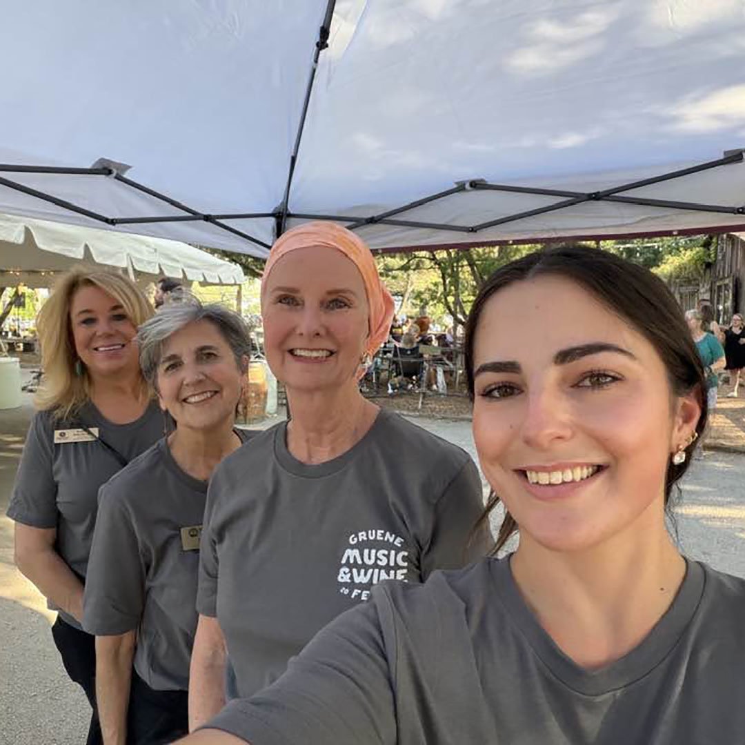 Smiling group of four women posing for a selfie under a tent at an outdoor event, all wearing gray shirts.”] ,